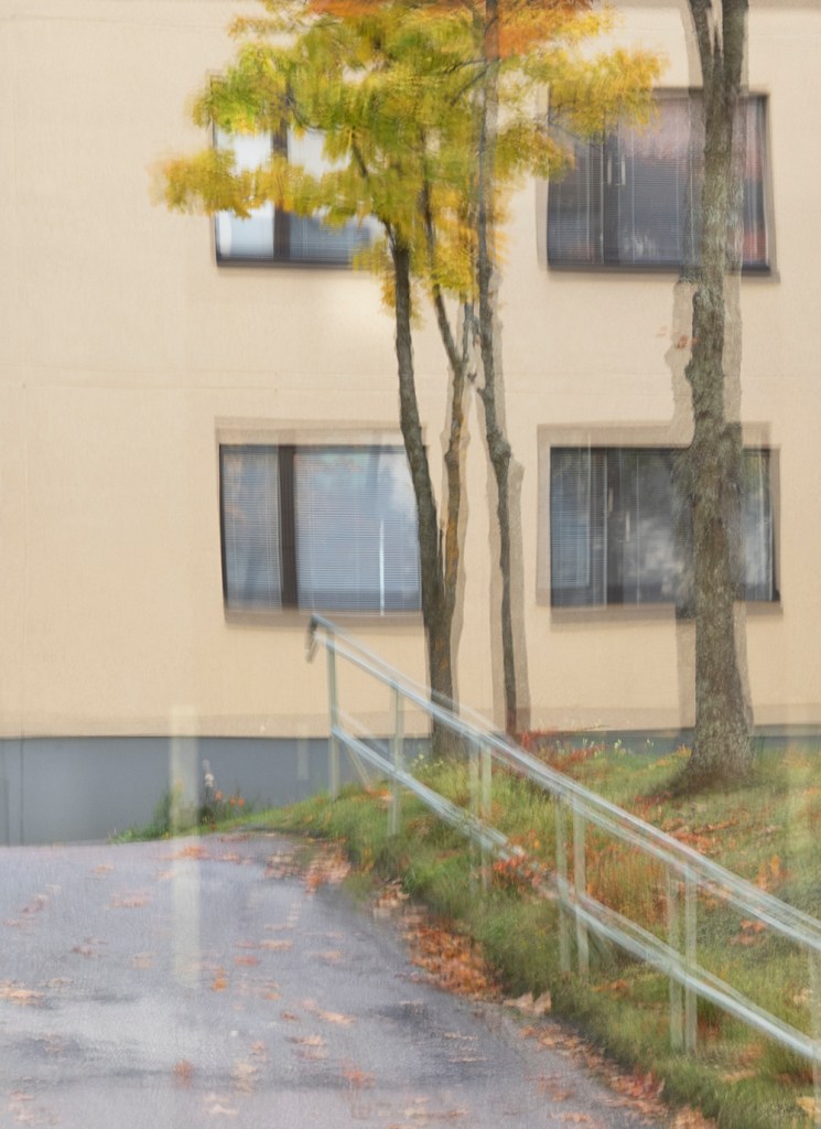 kerrostalon ikkunoita ja muutama puu näkyvät hieman epäselvänä heijastuksena toisesta ikkunasta