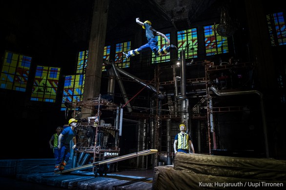 Sirkustaiteilijat ovat pukeutuneet rakennustyömiesten vaatteisiin ja taiteilevat keinulaudalla